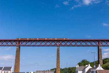 Queensferry, Scotland, UK - June 14, 2012: Train rides over Red metal iconic Forth Bridge over Firth of Forth against blue sky. Tall supporting pillars and rooftops of the town. 