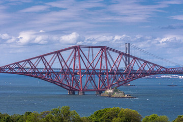 Queensferry, Scotland, UK - June 14, 2012: Closeup of One of three segments of Red metal iconic Forth Bridge over Firth of Forth between blue sky and water. Buildings on other side of water. 