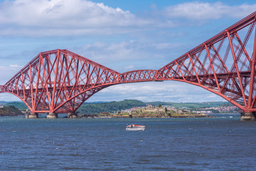 Queensferry, Scotland, UK - June 14, 2012: Segments of Red metal iconic Forth Bridge over Firth of Forth between blue sky and water. Brown buildings of old Battery on other side of water. Ferry boat.
