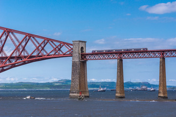 Queensferry, Scotland, UK - June 14, 2012: Train rides over Red metal iconic Forth Bridge over Firth of Forth against blue sky. Tall supporting pillars and some ships on the water.