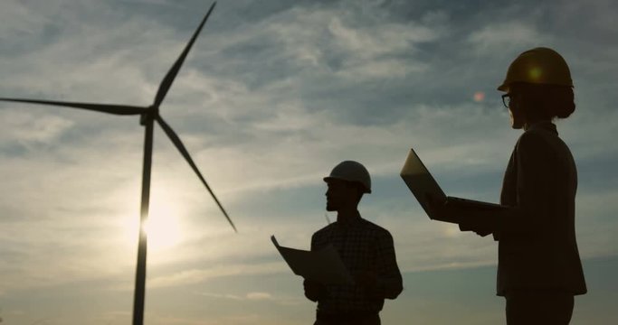 Male engineer in a helmet holding some documents and drawings while having a discussion with his female boss with a laptop computer at the windmills renewable energy station. Shadows in the twilight.