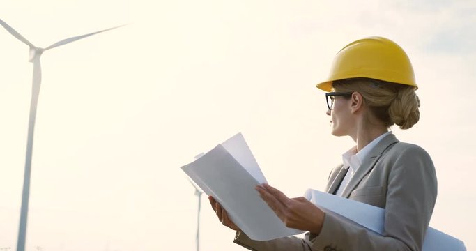 Portrait shot the woman in a helmet, environmental and safe power engineer, standing with some documents while watching the work of the big windmill turbine.