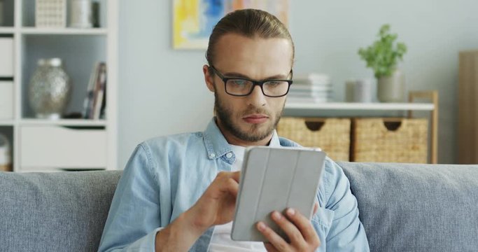 Serious attractive man in glasses using his tablet computer while sitting on the couch a home. Portrait shot.
