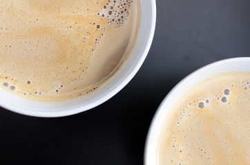 Close up of two cups of hot milk coffee with foam. Detail of cups on a black tray. Top view with focus on the surface of the coffee full of bubbles. Copy space.