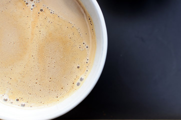 Close up of a cup of hot milk coffee with foam. Detail of cup on a black tray. Top view with focus on the surface of the coffee full of bubbles. Copy space.
