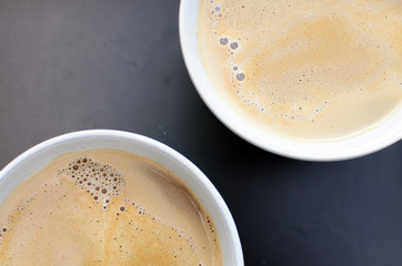 Close up of two cups of hot milk coffee with foam. Detail of cups on a black tray. Top view with focus on the surface of the coffee full of bubbles. Copy space.