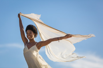Smiling Young Woman Wearing a White Dress and Holding a Blowing Scarf Against a Blue Sky