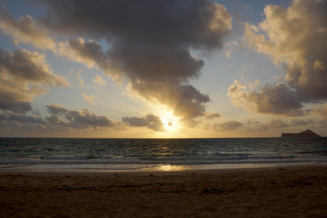 Fototapeta na wymiar Early Morning Sunrise on Waimanalo Beach over Rock Island bursting through the clouds