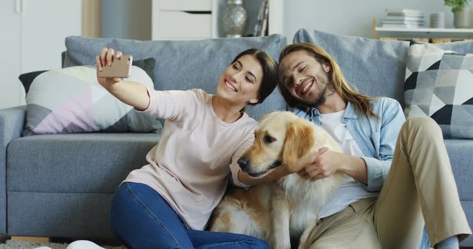 Young Caucasian couple sitting on the floor with a dog and leaning on a sofa, woman taking selfie photos on the smartphone. Portrait shot.