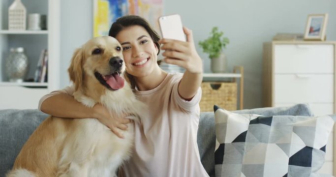Portrait shot of the charming brunette young woman sitting on the couch and hugging a labrador dog and taking selphie photos on the smartphone.
