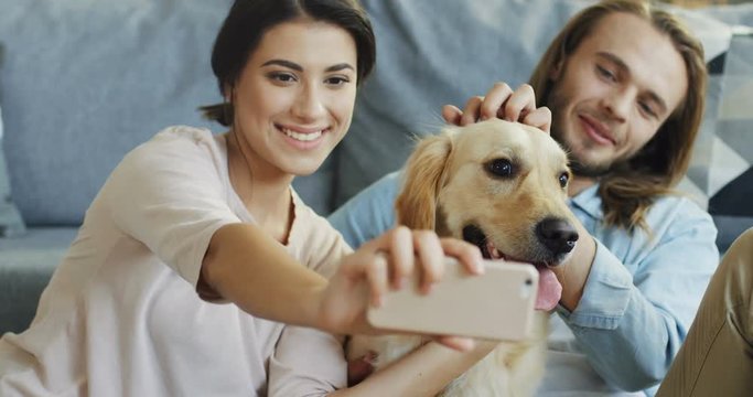 Close up of the happy smiled boyfriend and girlfriend hugging, kissing and petting their dog while taking selfie photos on the smartphone. Inside.