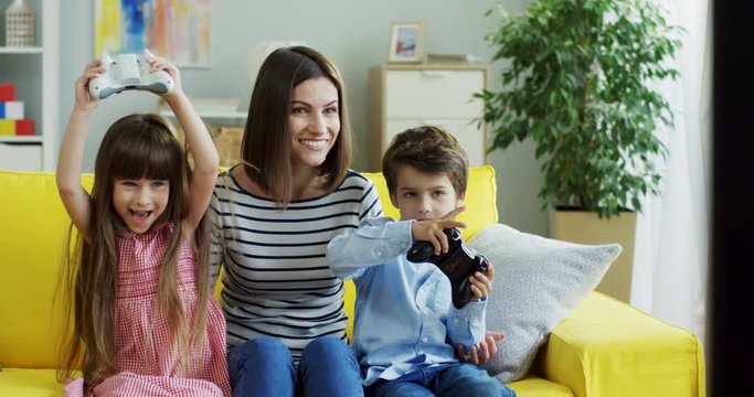 Cute little son and daughter playing videogame with joysticks while sitting near their mother and she hugging them. At home.