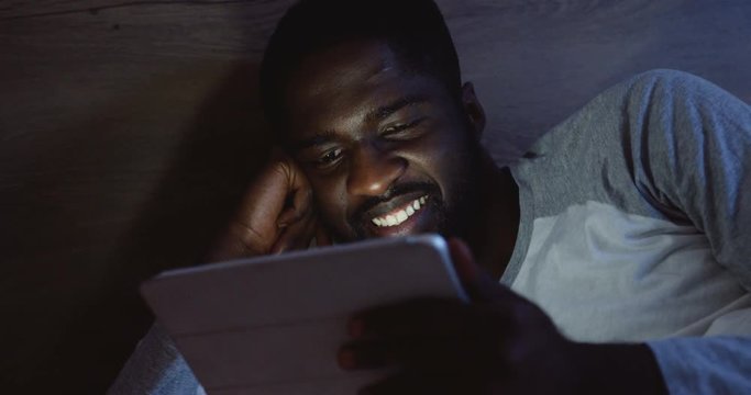 Close uop of the joyful young African American man with a tablet device in hands looking at the screen and laughing late at night.