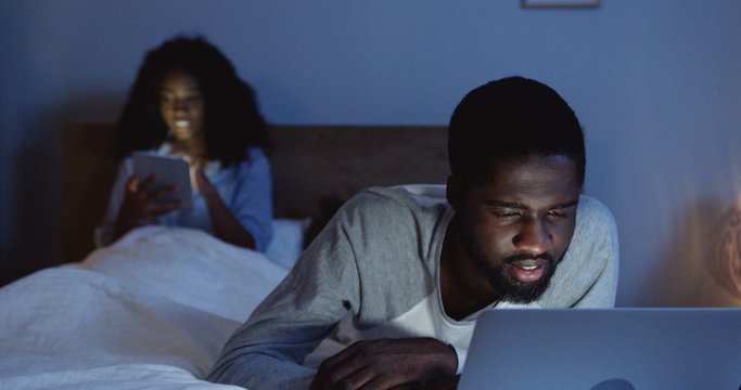 Good looking African American man lying on the bed in pajama and watching something funny on the laptop late at night. Her girlfriend with tablet computer sitting on the background and smiling.