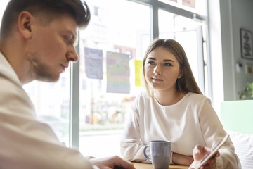Girl is sitting at table with man and holding phone in hand. She is showing something to him. He is looking seriously at phone. They are working together.