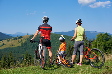 Back view of tourist family bikers, mom, dad and child standing with bikes on top of grassy hill,...