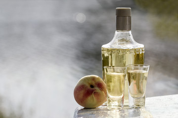 Bottle of tequila with glasses and a peach on  white table near lake