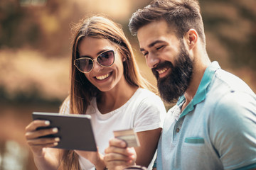 Cheerful young couple sitting on a park bench and uses a digital tablet for online shopping