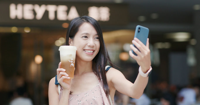 Woman holding a cup of cheese green tea and taking selfie on cellphone at Heytea store in the city