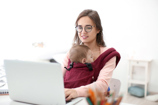 Young Woman With Baby In Ergo Carrier Using Laptop Indoors