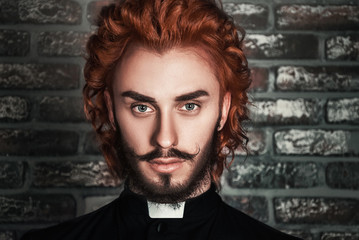 Close-up portrait of young red-haired man wearing priest garment, with designer stubble (on brick...