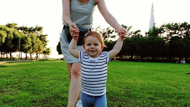 Young pretty mother with her Cute Little laughing baby daughter walking by grass at sunset in a park in suumer day. Slow Motion. Happy childhood. Love and Family