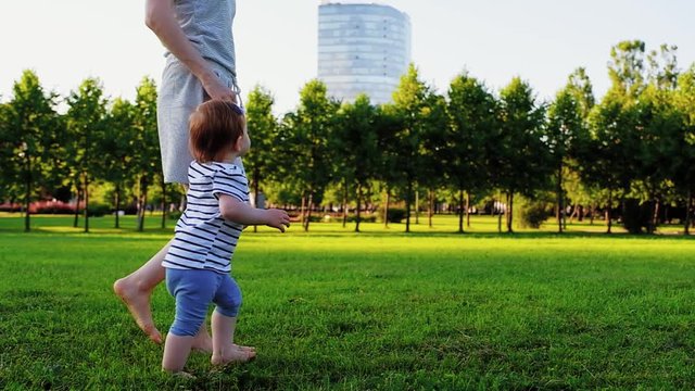 Young pretty mother with her Cute Little laughing baby daughter walking by grass at sunset in a park in suumer day. Slow Motion. Happy childhood. Love and Family