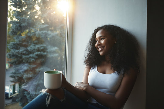 People, leisure, rest, relaxation and lifestyle concept. Picture of fashionable girl with wavy hair smiling happily, having nice time at home, sitting on windowsill and drinking herbal tea