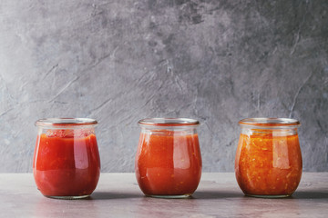 Variety of three homemade tomato sauces in glass jars on grey kitchen table. Close up