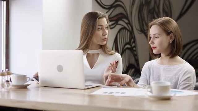 Two young business women are working with a laptop in a cafe. They are discussing and preparing the contract. It is a business meeting. Girls are sitting in the cafe at the table and messaging on