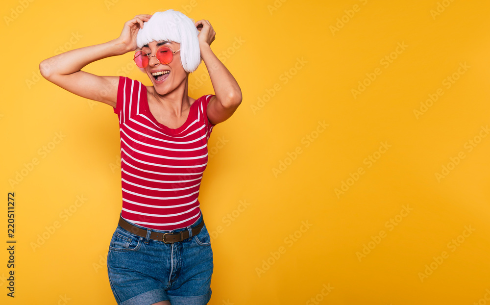 Wall mural Beautiful young woman in blonde wig and pink sunglasses having fun on orange background, fashion blonde posing