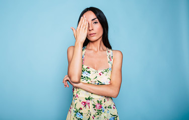 Young attractive brunette woman closed one eye with help of her hand on blue background