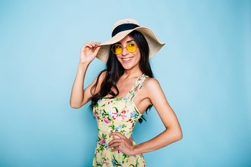 Portrait of cute summer brunette woman in hat, sunglasses and colorful dress, stylish girl have a fun and posing on blue background