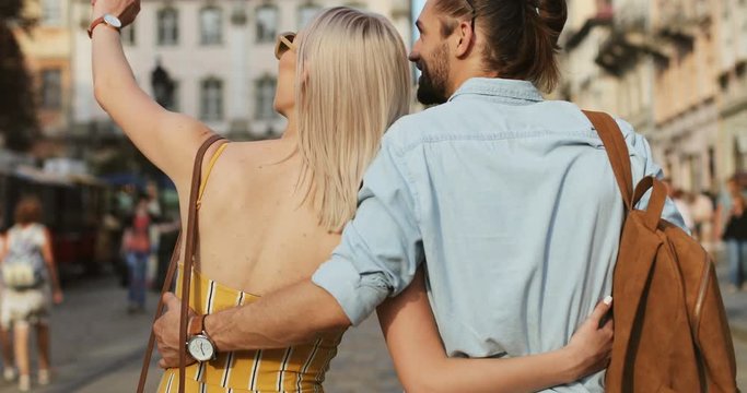 Back view on the young Caucasian couple hugging and walking a town square while coming there as tourists. Rear.