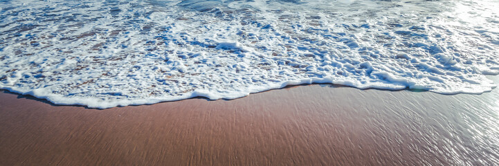white foamy oceanic sea wave runs over a sandy beach, natural abstract background and texture, panorama banner format