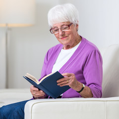 Portrait of a senior woman at home - Looking happy, looking at the camera, smiling while sitting on the sofa in her living room and reading a good book