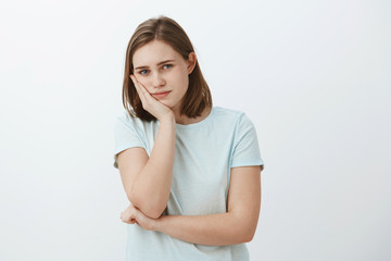 Girl feeling bored from routine. Portrait of calm good-looking european female leaning head on palm gazing at camera with tiresome and indifferent look having nothing to do standing against grey wall