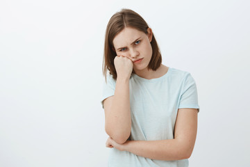 Fototapeta premium Studio shot of displeased offended and gloomy cute female brunette in t-shirt leaning face on fist frowning and pursing lips feeling envy or jealous seing sibling received awesome gift from parents