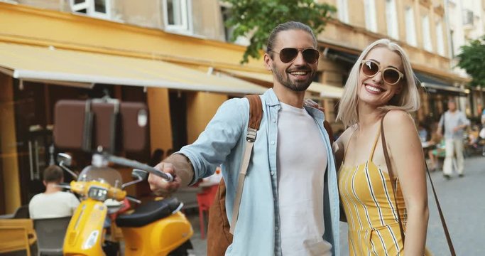 Young attractive Caucasian man and woman taking photos on the smartphone with selfie stick. blowing kisses and hugging on the street.