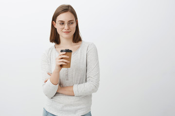 Wow nice butt. Portrait of naugty good-looking enthusiastic woman with brown hair in glasses smirking and gazing at lower right corner drinking coffee while standing near cafe posing over gray wall