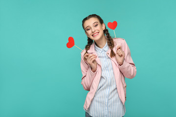 Happy beautiful girl in casual style, pigtail hairstyle and pink jacket, standing and holding red heart stickers and looking at camera and toothy smiling, Indoor, isolated on blue or green background