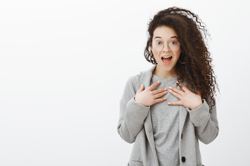 Portrait of surprised thrilled girlfriend with curly hair in trendy transparent glasses and grey coat, holding palms on chest, gasping and smiling broadly with opened mouth, feeling excited