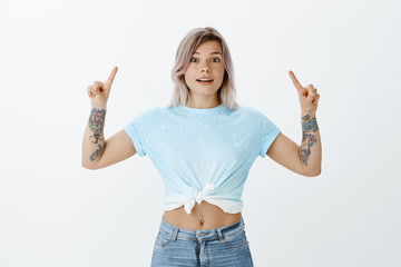 Indoor shot of happy carefree european female with fair hair and tatoos, smiling broadly and pointing up with index fingers, being amazed and interested in new copy space over grey wall