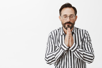 Never forgets to thank god for success. Calm and relaxed good-looking believer in glasses and striped shirt, holding hands in pray while praying or meditating with closed eyes over grey wall