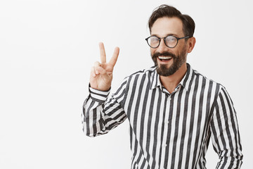 Fototapeta premium Peace. Attractive adult man with beard and moustache in glasses and formal outfit trying to find contact with youngsters, showing victory sign and smiling broadly while talking with friend of son