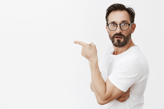 Is it him. Portrait of shocked and curious good-looking mature guy in glasses and t-shirt, standing in profile and turning to camera asking question, pointing left with index finger, being interested