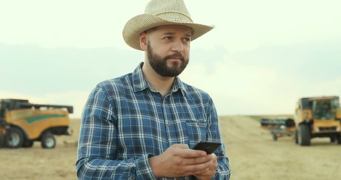Caucasian young farmer in the hat scrolling, taping and typing on the smartphone while the harvest being gathered.