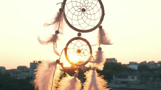 Amulet dreams catcher waving in a light wind against the backdrop of a bright sunset and a bokeh of city lights. White light feathers rise in the wind. The disc of the setting sun. Ethnic amulet