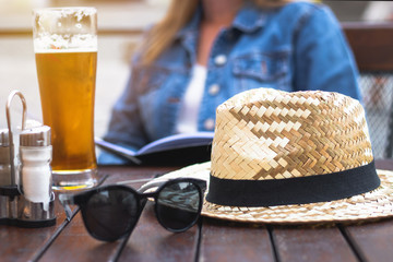 Woman is ordering food in outdoor restaurant. Straw hat and sunglasses on a wooden table. Glass of beer in pub.