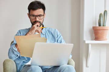Serious concentrated businessman in spectacles for vision protection, works on laptop computer,...
